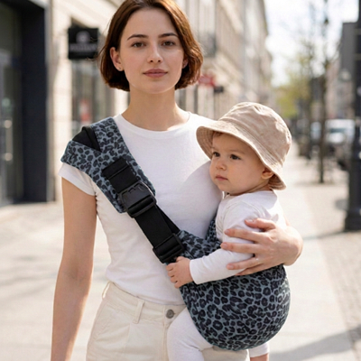  Maman soutenant bébé coiffé d’un chapeau beige dans l’écharpe de portage Harmonie à motif léopard gris, lors d’une sortie en ville.