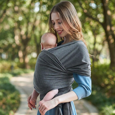 Maman portant son bébé contre elle dans une écharpe de portage Nomade grise sur un sentier arboré.