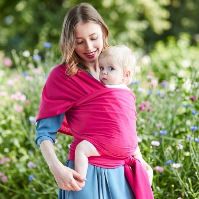 Maman en robe bleue portant son enfant dans une écharpe de portage Nomade rose dans un jardin fleuri.