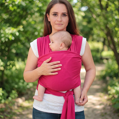 Maman portant son bébé contre elle dans une écharpe de portage Nomade rose, dans un parc.