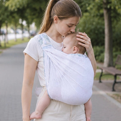 Maman porte son bébé dans une écharpe de portage Pastel blanche au parc.
