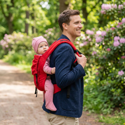 Papa dans un parc avec bébé au dos dans un porte-bébé 3 en 1 rouge